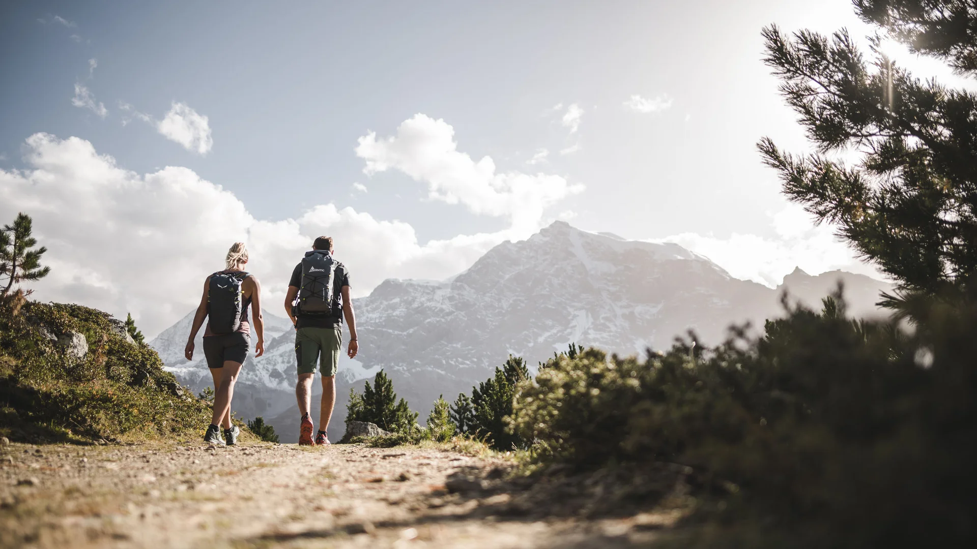 Zwei Wanderer mit Rucksäcken auf Bergweg vor schneebedeckten Gipfeln