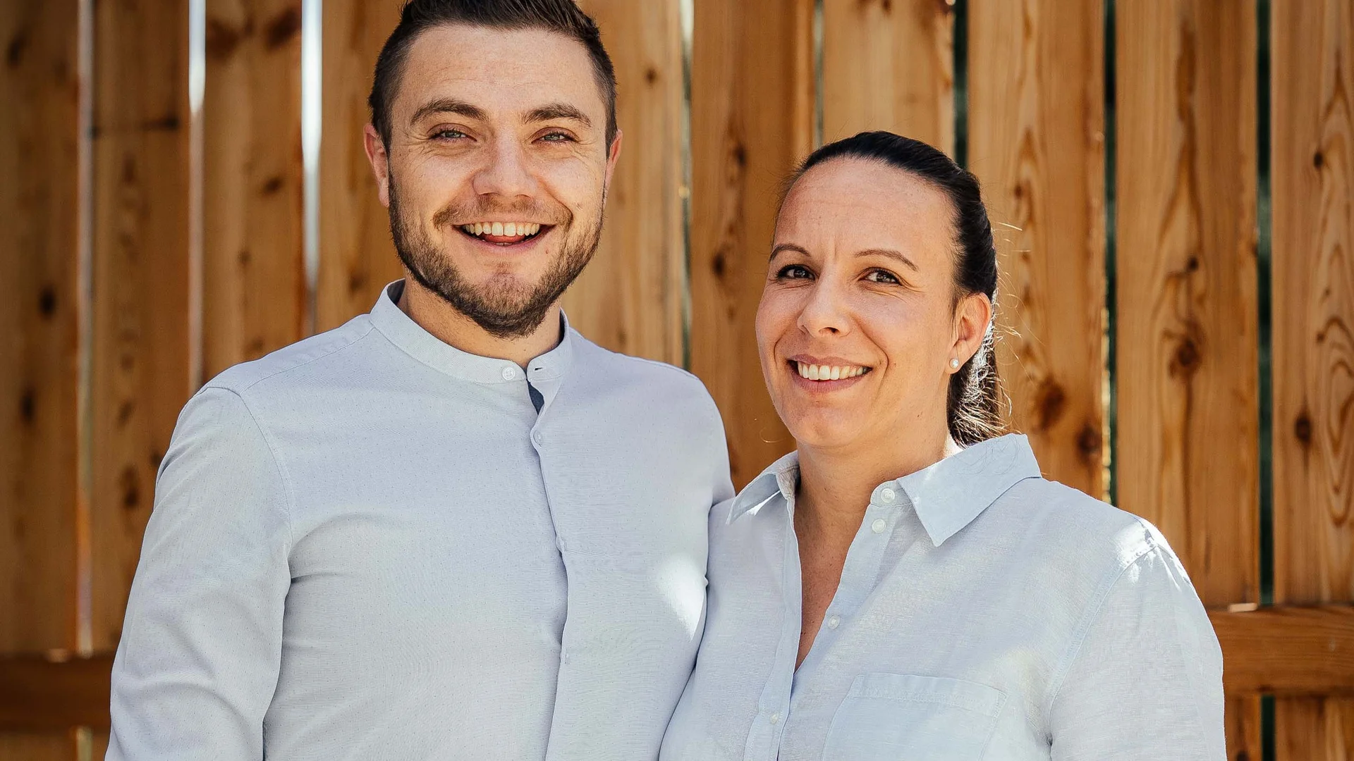 Uomo e donna sorridenti con camicie bianche davanti a parete di legno