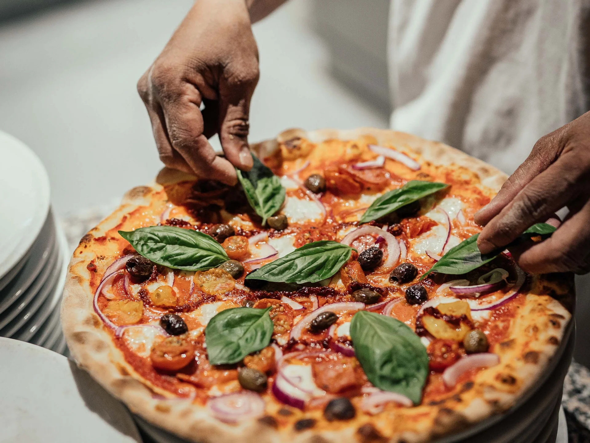 Der Lodenwirt in Vintl | Hotel, Residence & Restaurant Hände legen frische Basilikumblätter auf eine Pizza mit Tomaten und Oliven
