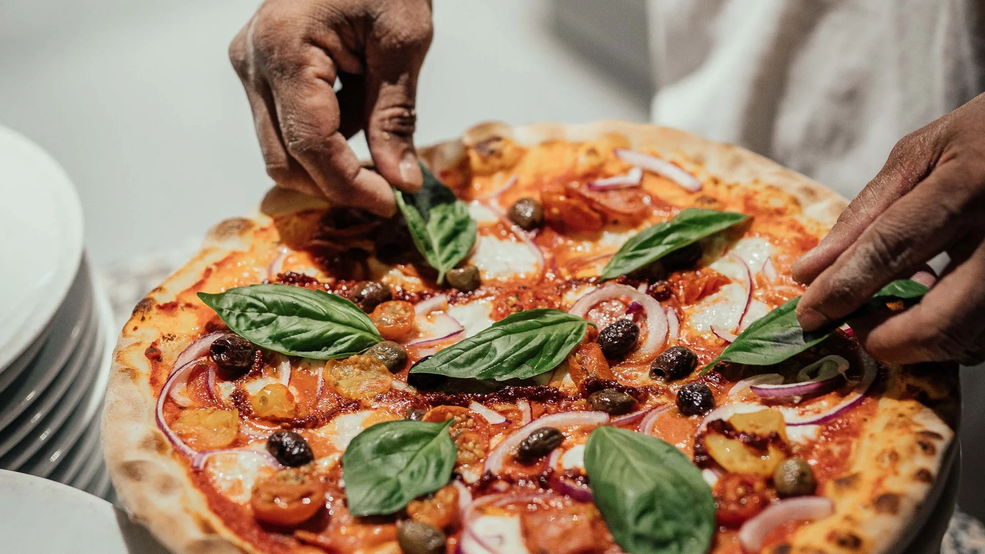 Mani che posizionano foglie di basilico fresco su una pizza con pomodori e olive