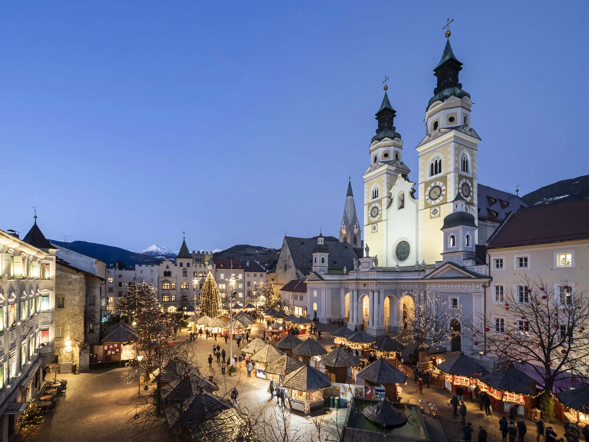 Il Lodenwirt a Vandoies | Hotel, residence e ristorante Mercatino di Natale illuminato in una piazza davanti a una chiesa con due torri al crepuscolo
