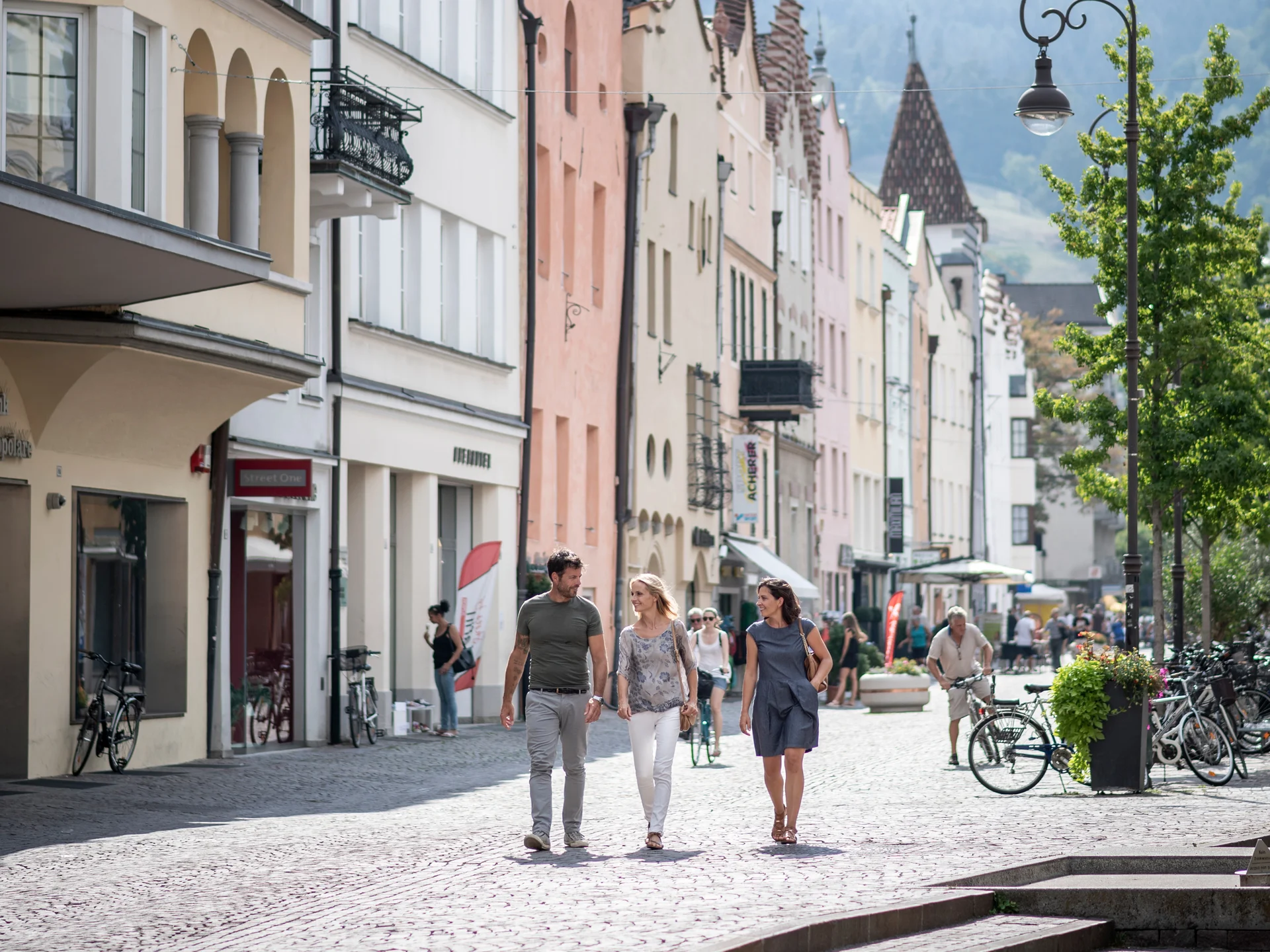 Der Lodenwirt in Vintl | Hotel, Residence & Restaurant Menschen spazieren in einer Fußgängerzone mit bunten Häusern und Bergen im Hintergrund
