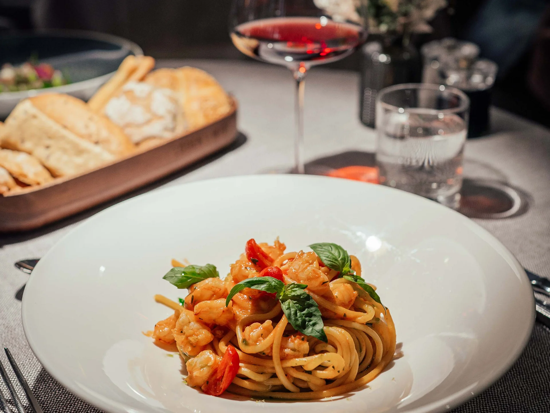 Piatto di pasta con gamberi, bicchiere di vino rosso e cestino di pane su tavolo apparecchiato