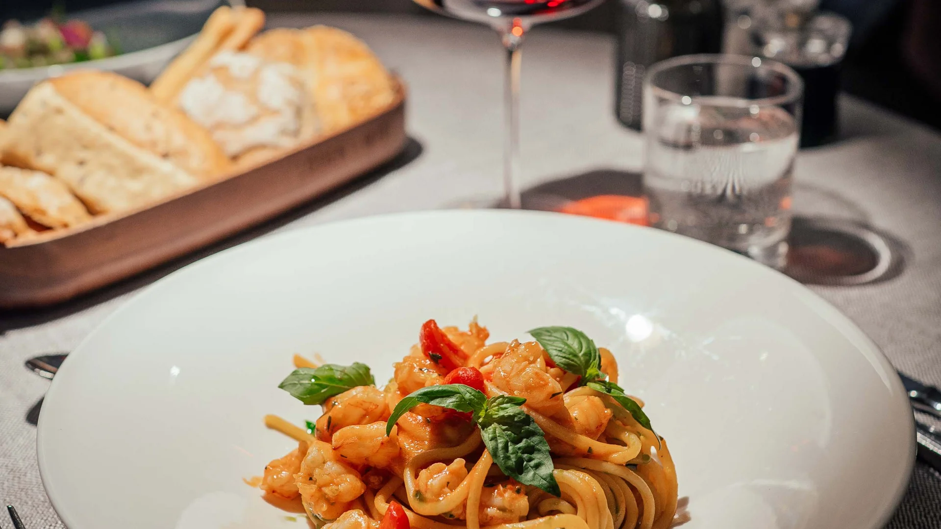 Piatto di pasta con gamberi, bicchiere di vino rosso e cestino di pane su tavolo apparecchiato