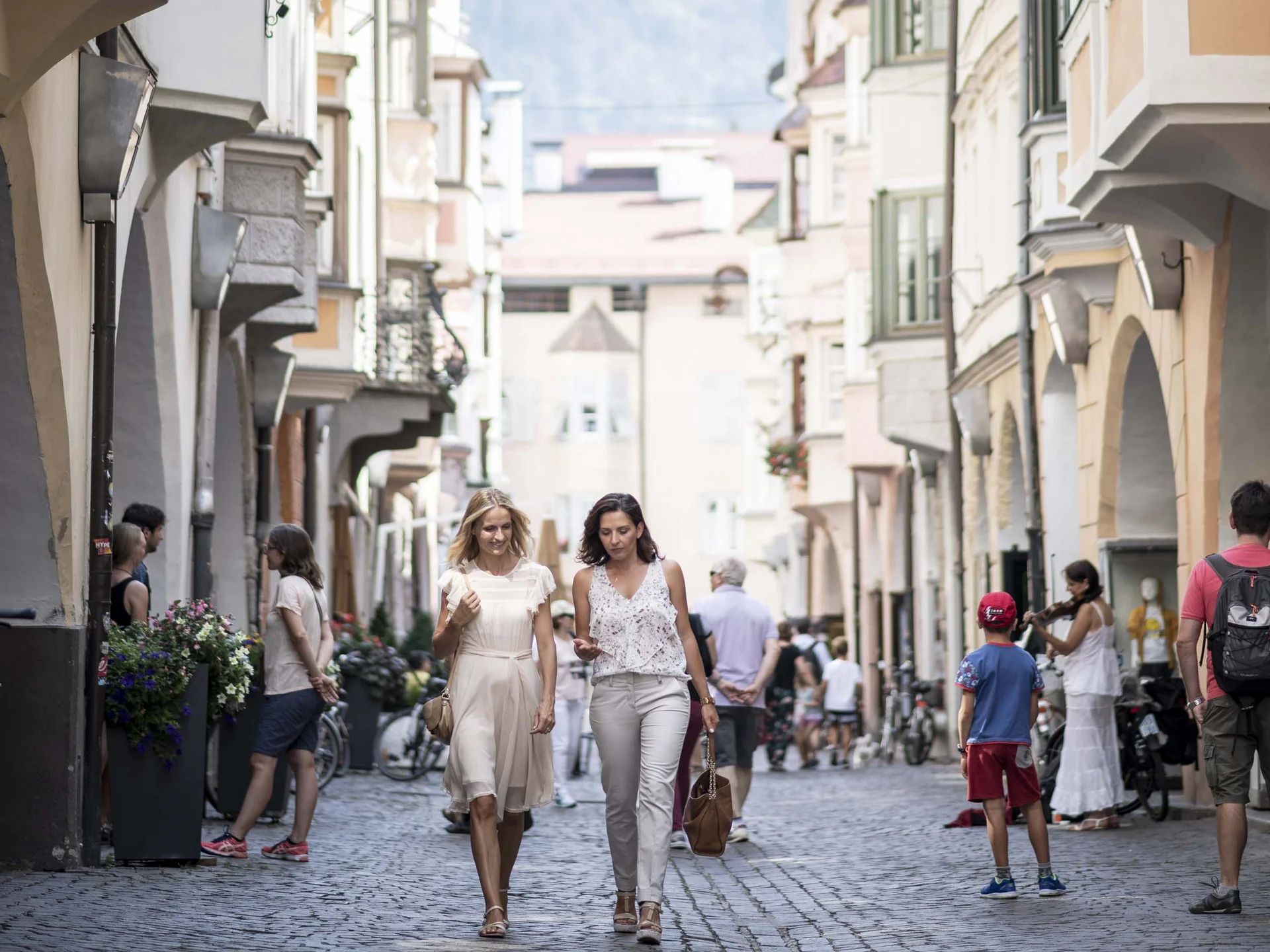 Le vostre vacanze in Val Pusteria Persone in una strada lastricata del centro storico con edifici storici e montagne sullo sfondo