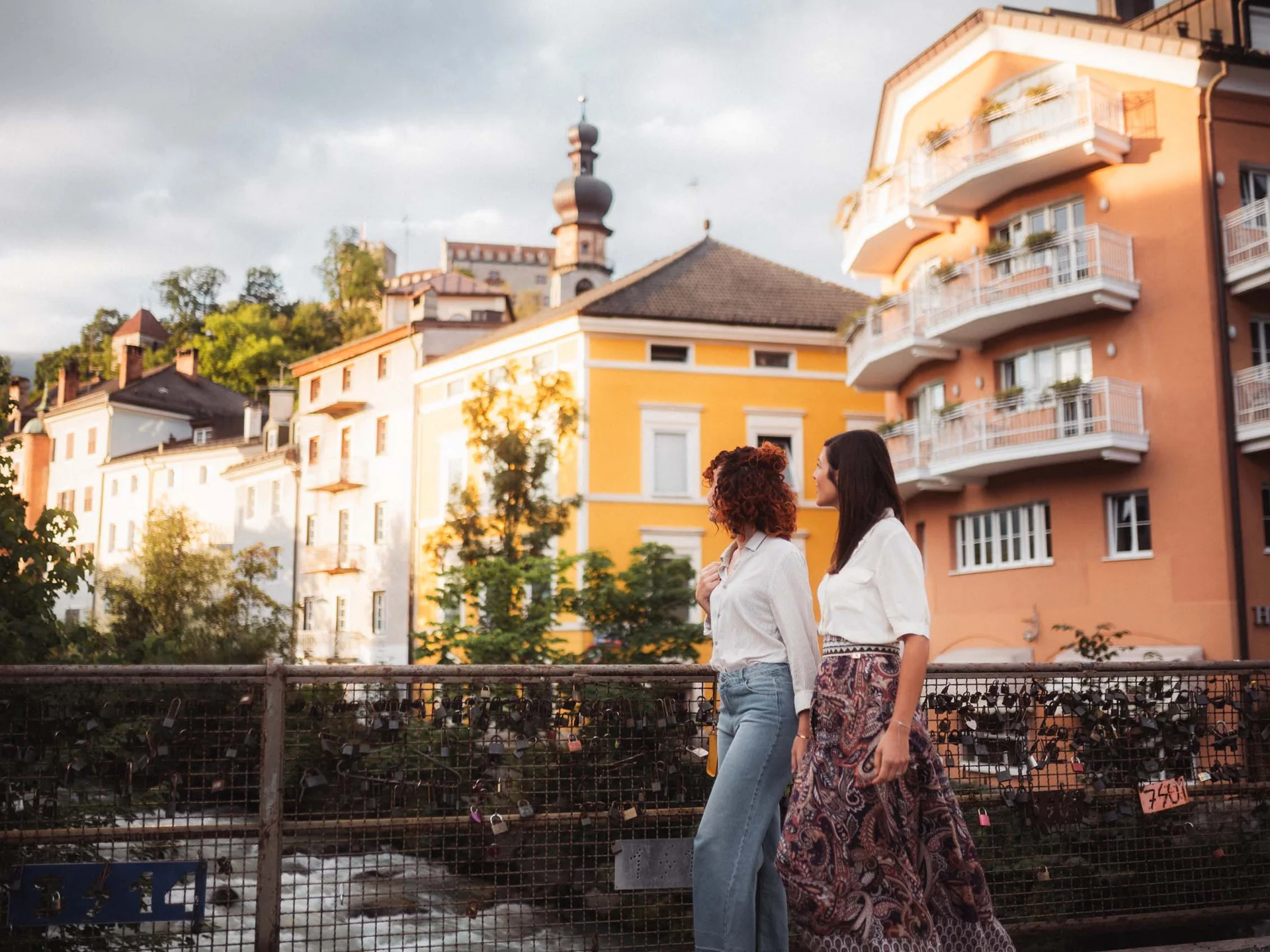 Le vostre vacanze in Val Pusteria Due donne camminano su un ponte davanti a edifici colorati e a un campanile