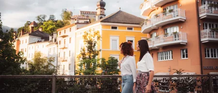 Due donne camminano su un ponte davanti a edifici colorati e a un campanile