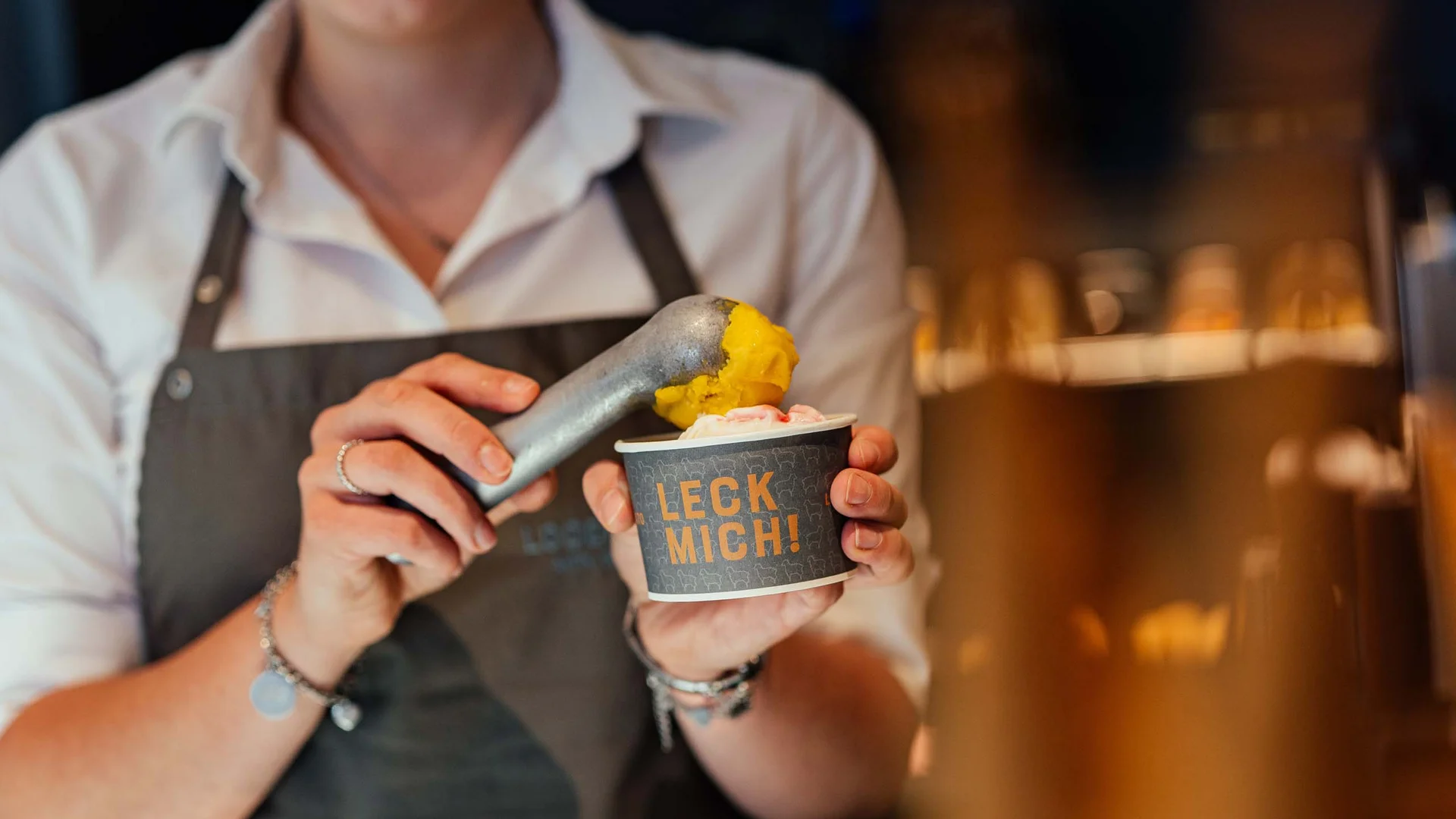 Persona con grembiule che serve gelato in coppetta con scritta Leck Mich