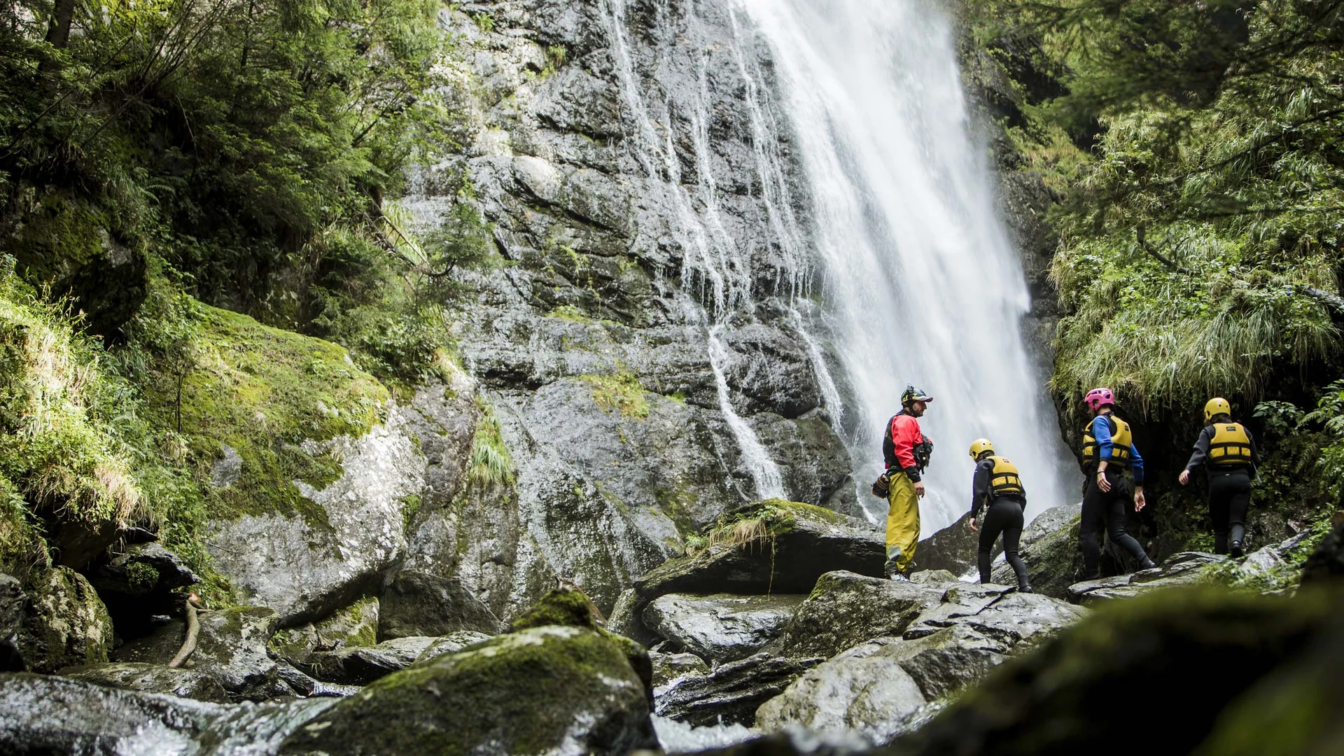 Quattro persone davanti a una cascata in una zona rocciosa e boscosa