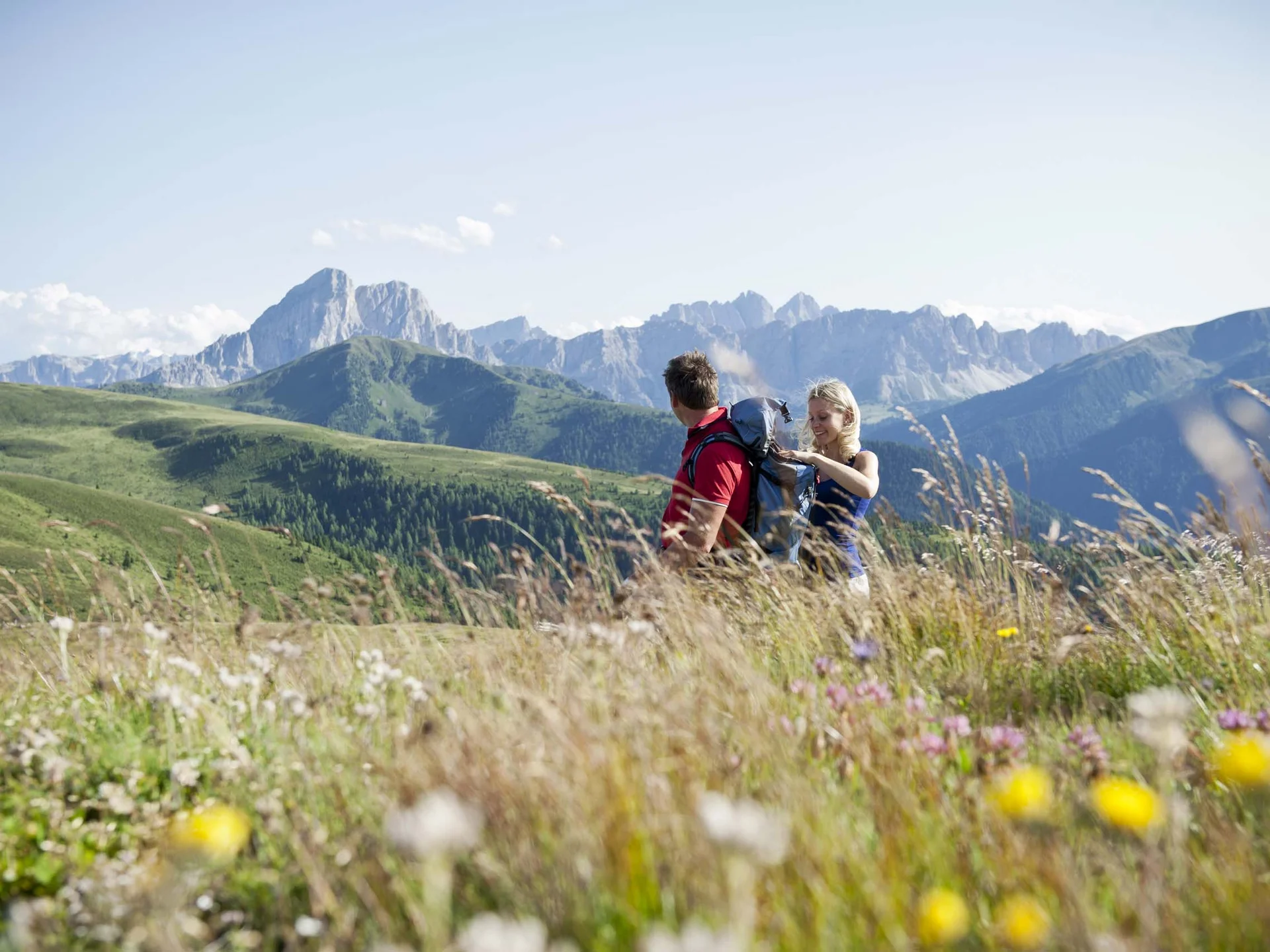 Le vostre vacanze in Val Pusteria Due escursionisti con zaini in un prato fiorito con montagne alpine sullo sfondo