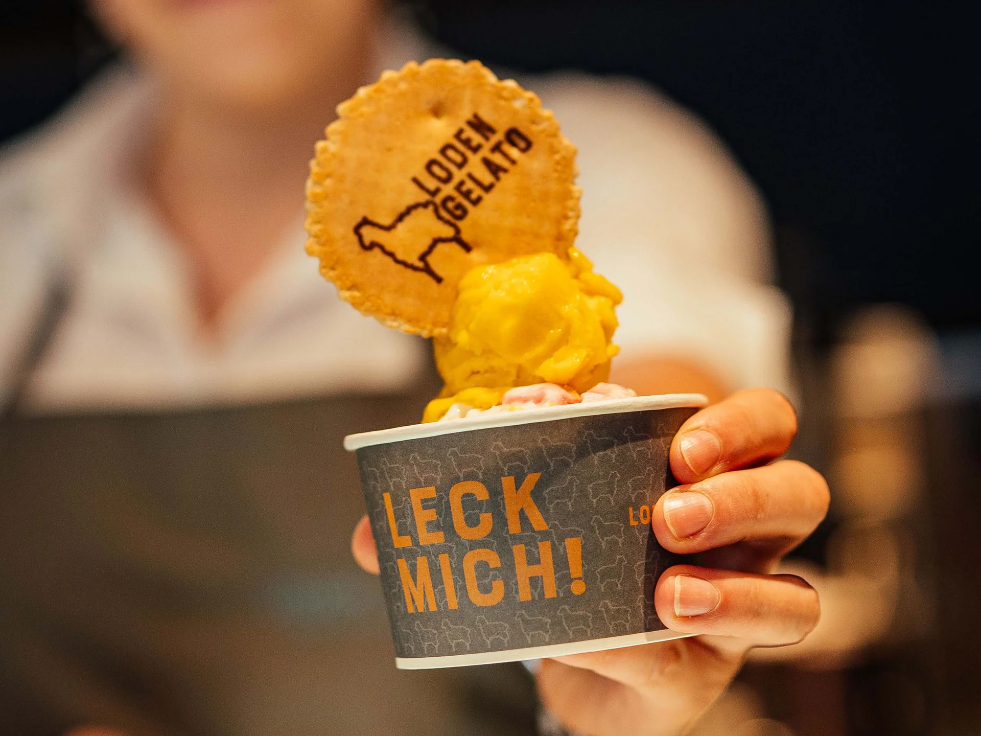 Persona che tiene una coppetta di gelato giallo con un biscotto con scritto Loden Gelato