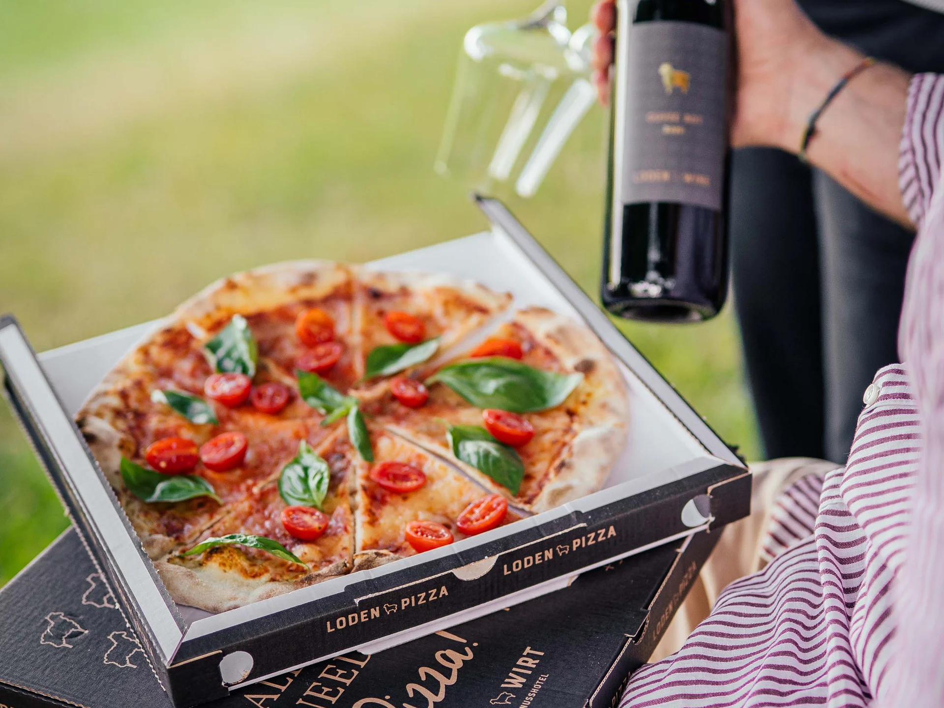 Der Lodenwirt in Vintl | Hotel, Residence & Restaurant Person hält Pizza und Weinflasche mit zwei Weingläsern im Freien
