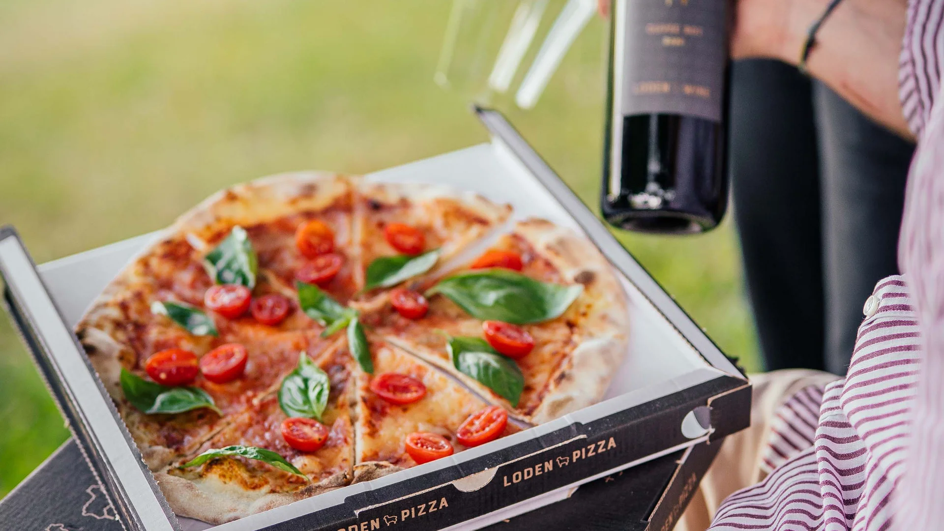 Persona tiene pizza e bottiglia di vino con due bicchieri da vino all'aperto