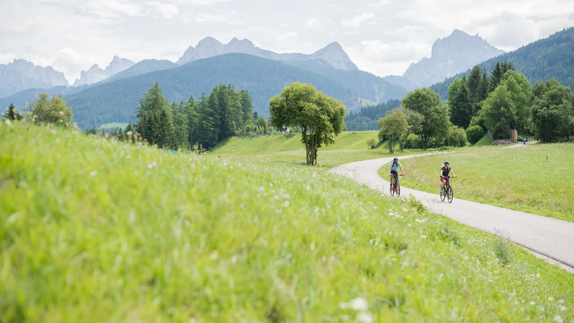 Zwei Radfahrer auf einem Weg durch grüne Wiesen mit Bergen im Hintergrund
