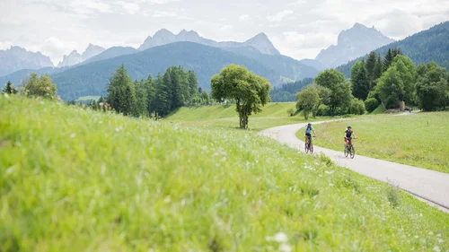 Offerte in Val Pusteria | Lodenwirt Vandoies Zwei Radfahrer auf einem Weg durch grüne Wiesen mit Bergen im Hintergrund