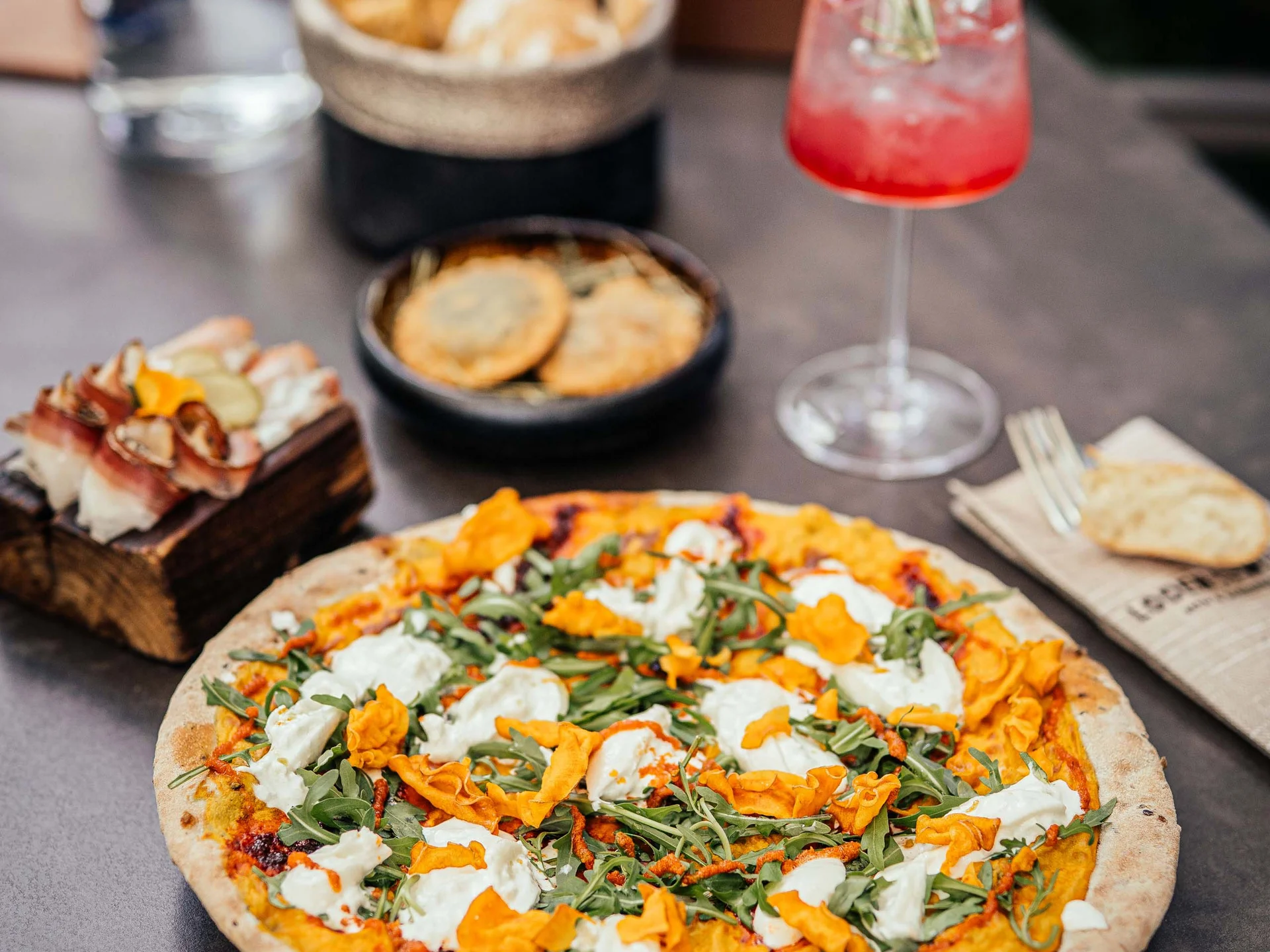Der Lodenwirt in Vintl | Hotel, Residence & Restaurant Pizza mit Rucola, Käse und Kürbisblüten auf einem Tisch mit Getränken und Snacks
