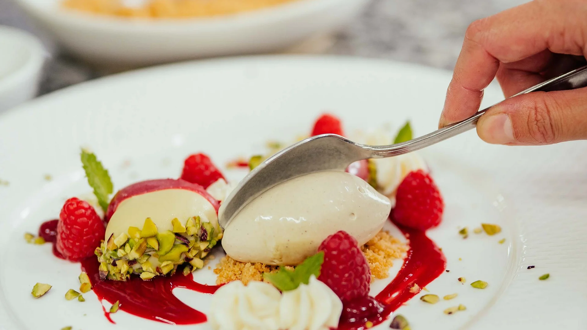 Mano con cucchiaio sopra dessert con lamponi, pistacchi e panna su piatto bianco
