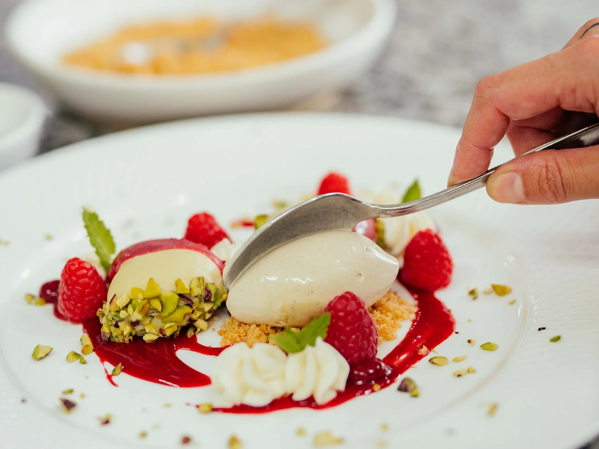 Der Lodenwirt in Vintl | Hotel, Residence & Restaurant Hand mit Löffel über Dessert mit Himbeeren, Pistazien und Sahne auf weißem Teller