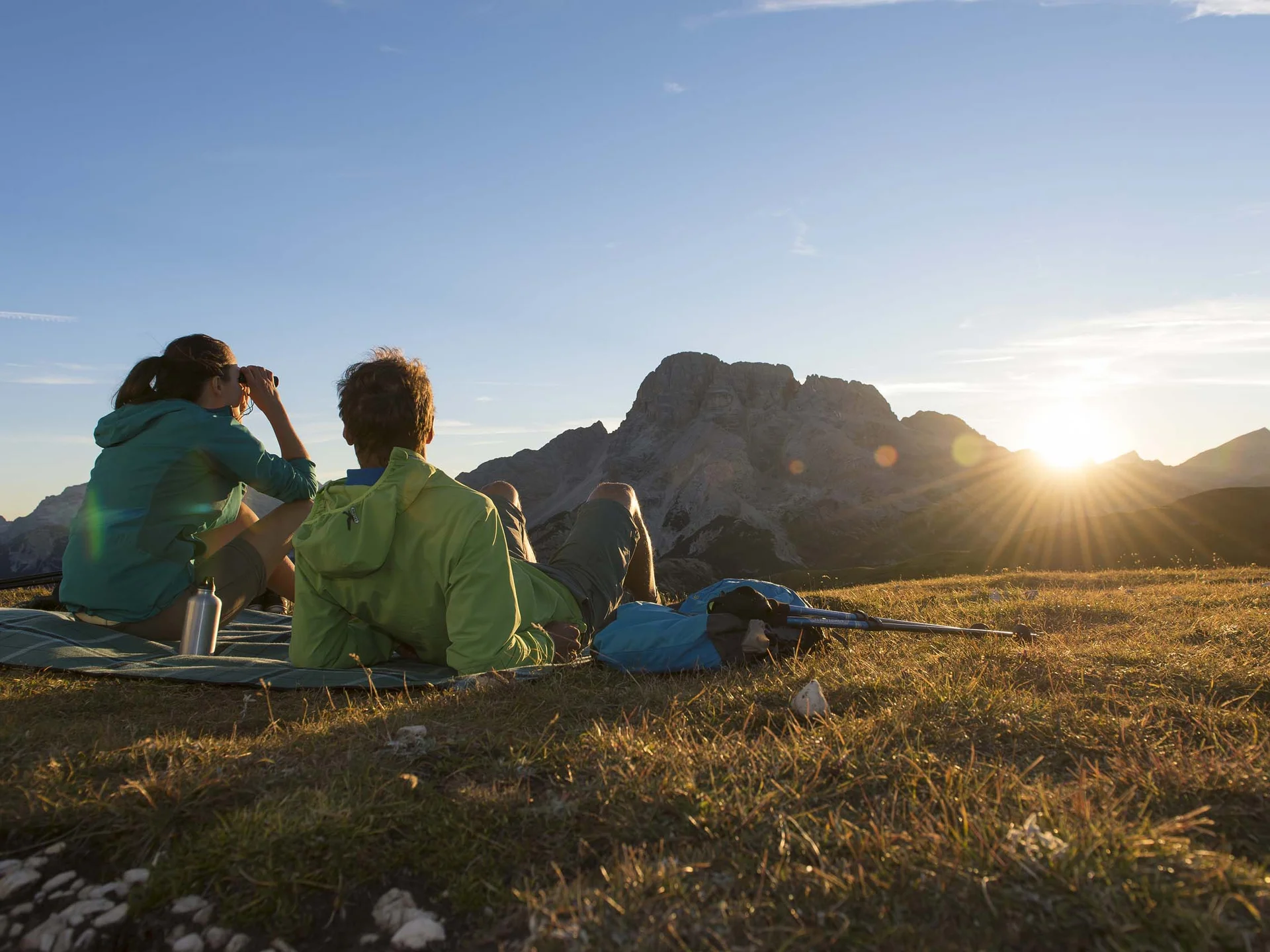 Der Lodenwirt in Vintl | Hotel, Residence & Restaurant Zwei Wanderer sitzen auf einer Wiese und schauen bei Sonnenuntergang auf Berge