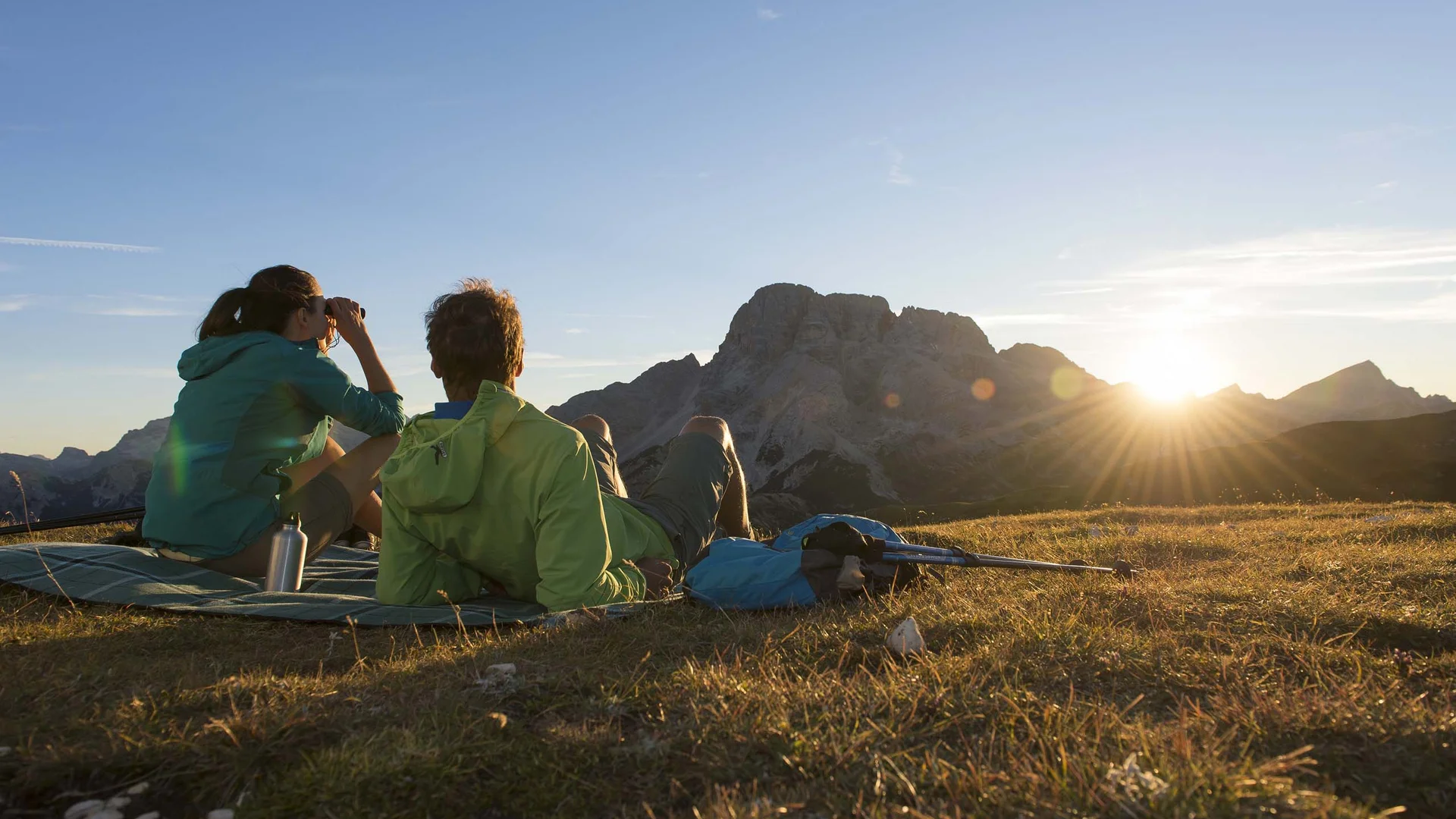 Due escursionisti seduti su un prato guardano le montagne al tramonto