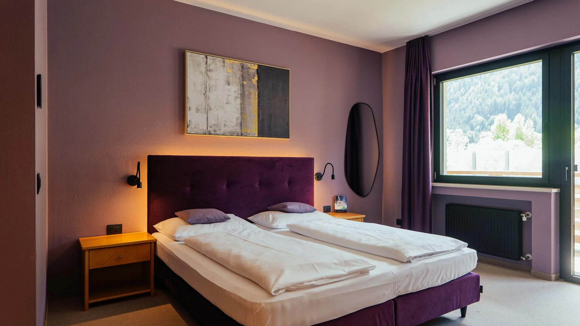 Modernes Hotelzimmer mit Doppelbett, lila Kopfteil und Blick auf Berge