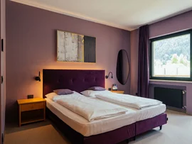 Der Lodenwirt in Vintl | Hotel, Residence & Restaurant Modernes Hotelzimmer mit Doppelbett, lila Kopfteil und Blick auf Berge