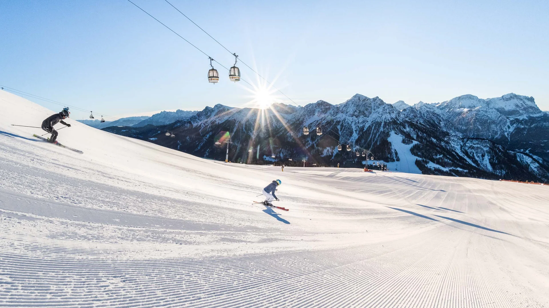 Hotel & Residence Lodenwirt photo gallery Skiers on a groomed slope with snowy mountains and cable cars