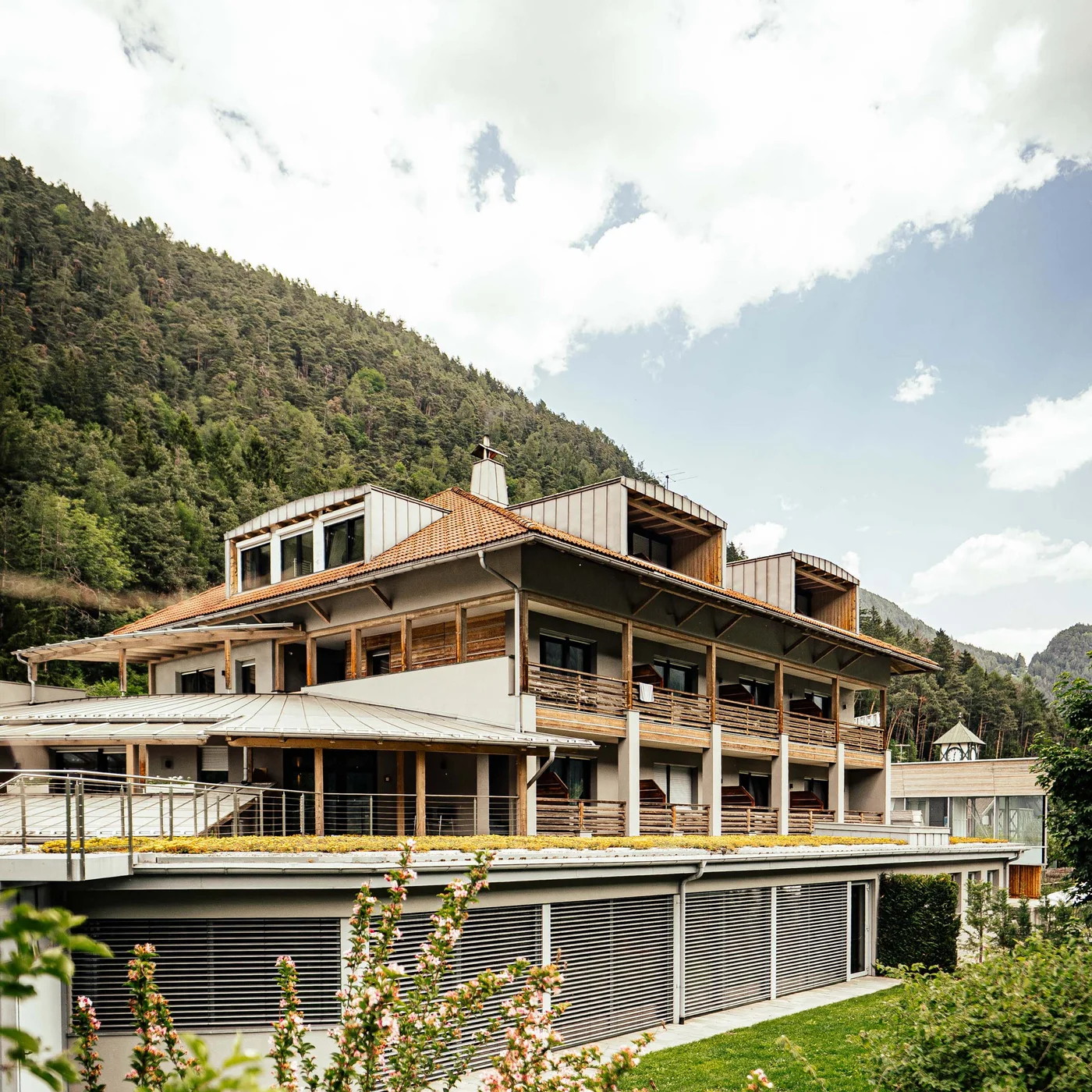 Der Lodenwirt in Vintl | Hotel, Residence & Restaurant Modernes Hotelgebäude vor bewaldetem Berg und bewölktem Himmel