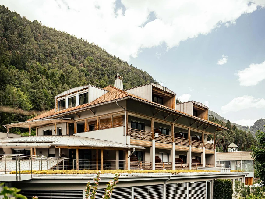 Der Lodenwirt in Vintl | Hotel, Residence & Restaurant Modernes Hotelgebäude vor bewaldetem Berg und bewölktem Himmel