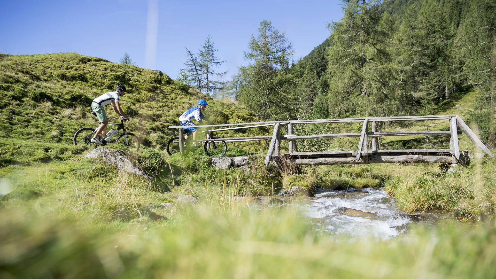 Hotel & Residence Lodenwirt photo gallery Two cyclists crossing a small wooden bridge over a stream in the mountains