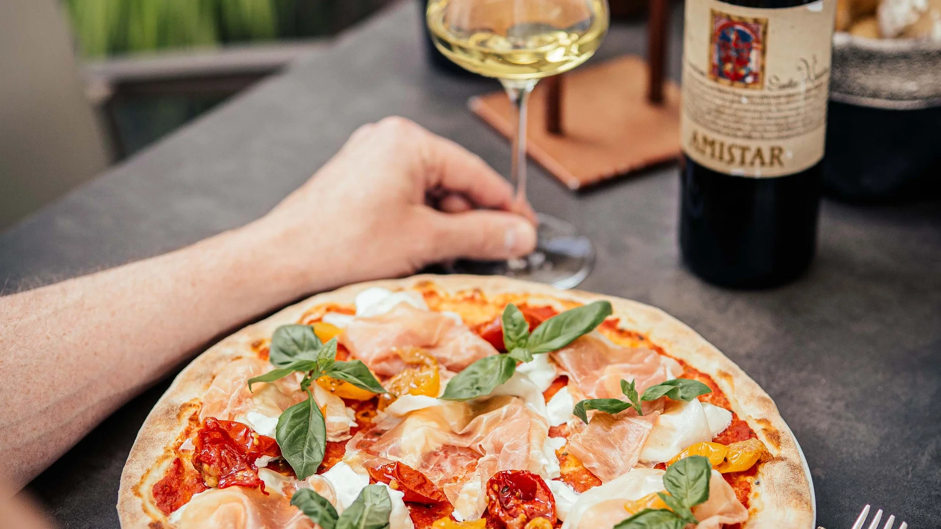 Pizza con prosciutto, basilico e pomodori, bicchiere di vino bianco e bottiglia di vino