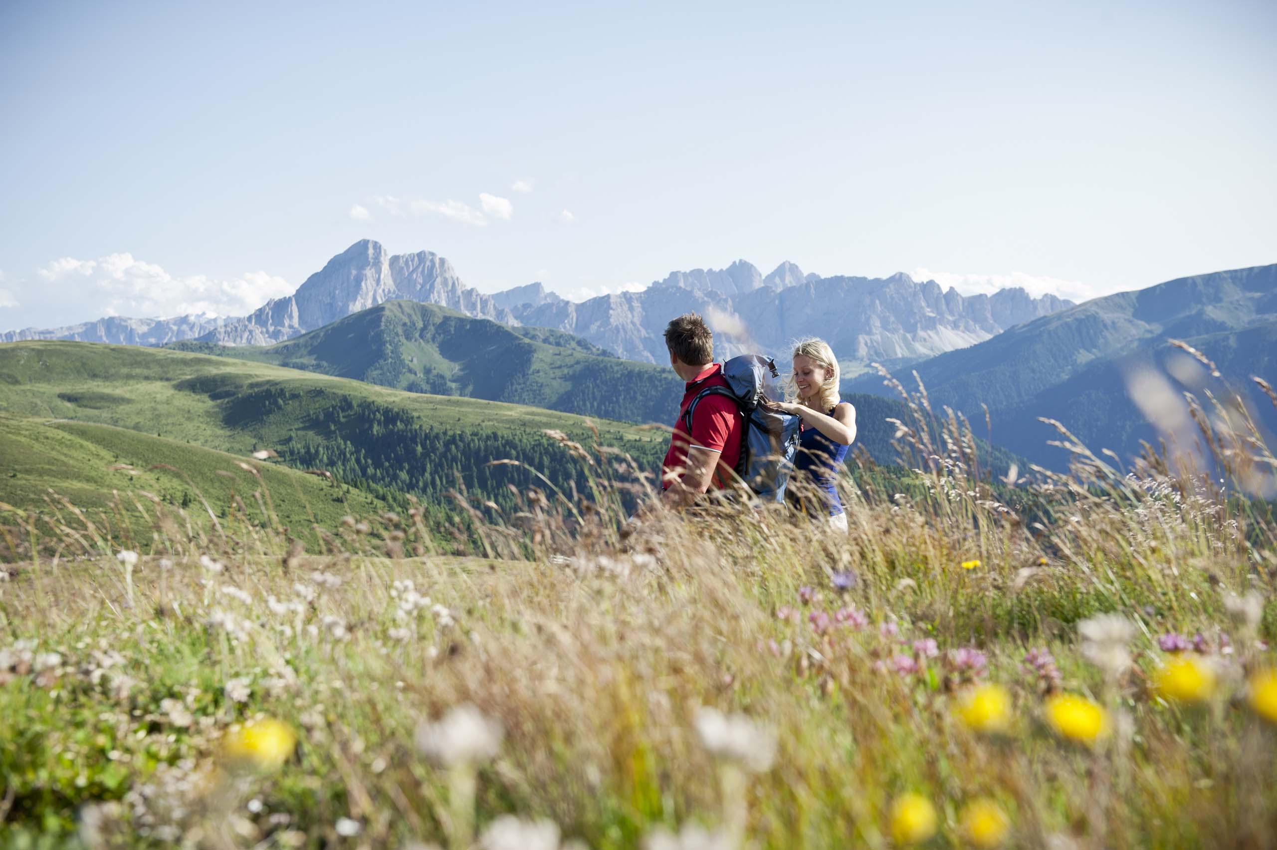 Due escursionisti con zaini in un prato fiorito con montagne alpine sullo sfondo