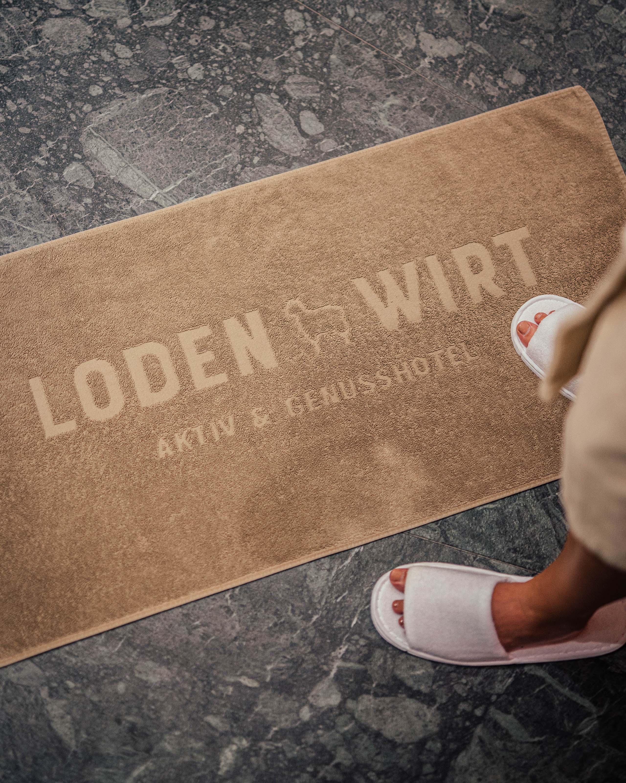 Additionals Person mit weißen Hausschuhen steht auf brauner Loden Wirt Badematte